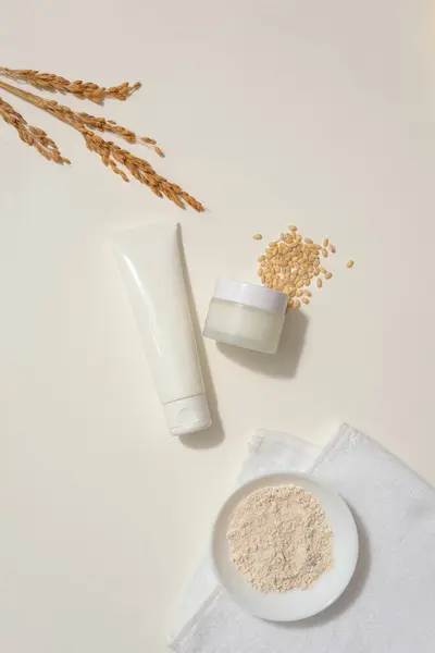 Flat lay of unlabeled jar and tube decorated with a handful of wheat seeds and wheat ears. A dish continuing rice bran placed on white towel. Mockup design with blank label containers