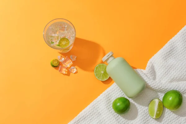 一个没有品牌的化妆品瓶子在白色面料的橙色背景下脱颖而出 旁边放着一杯加冰和新鲜柠檬的水 用天然精华制成的化妆品 — 图库照片
