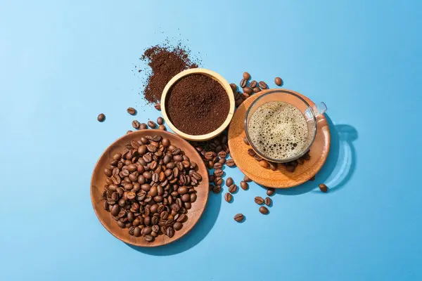 Coffee beans on a wooden plate, coffee powder in a wooden bowl and a cup of black coffee standing out on a blue background. Coffee brings many benefits to the body.