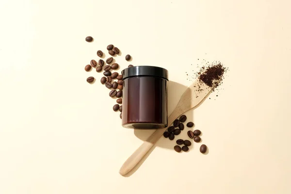 A brown scrub jar stands out against a pastel pink background with coffee beans. Using coffee powder to exfoliate the body provides the skin with essential nutrients, helping the skin stay healthy.