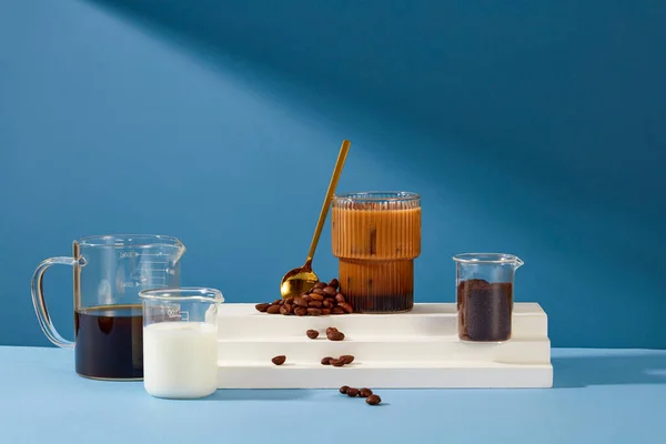 A glass of milky coffee is placed on a white platform with a metal spoon and coffee beans. Black coffee, fresh milk and coffee powder are stored in a beaker. Blue background for advertising.