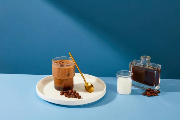 A cup of milk coffee is placed on a ceramic plate with a metal spoon and coffee beans, a cup of milk and a bottle of black coffee standing out against the blue background.