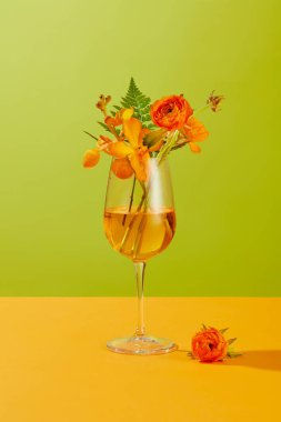 Taze çiçekler ve yeşil yapraklar, içinde açık sarı bir sıvı bulunan bir şarap bardağına konur. Boş alanla yaratıcı ol. Yeşil-sarı arkaplan