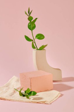 Yeşil yapraklar, bir seramik vazo, bir taş platform ve bir bez havlu açık pembe arka planda yan yana yerleştirilir. Ürün gösterimi için boş podyum.
