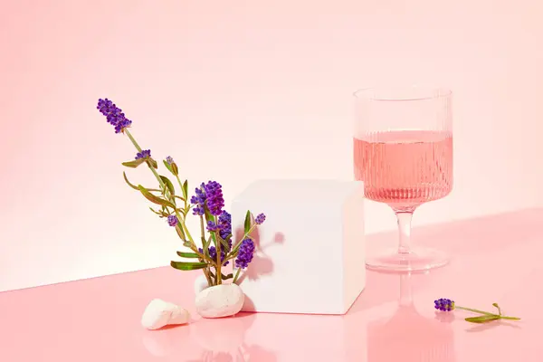 On a light pink background, a white podium and a wine glass with pink liquid create an inviting display, providing ample space for showcasing the beauty derived from lavender flowers.