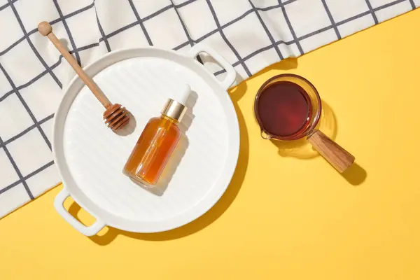 A bottle of serum and a honey drizzle on a white ceramic tray, honey in a glassware and a checkered tablecloth on a yellow background. Copy space for ads.