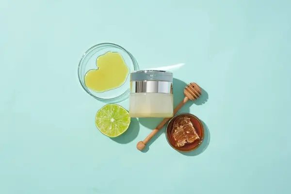 Honey in a petri dish, a luxury cosmetic jar, a honey drizzle and lemon are displayed on a pastel background. Honey helps balance bacteria on the skin, helping to protect the skin from infection.