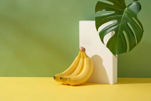 白い色の長方形の表彰台に傾いているバナナの束が付いている最小の場面 バナナ エキスは 老化肌を滑らかにしながらアクネの傷やダークスポットをフェードするのに役立ちます — ストック写真