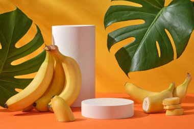 Sarı arka planda muz ve yeşil canavar yapraklarıyla süslenmiş beyaz podyumların önü. Kozmetik ürünlerinin sunumu için boş alan - muz özü