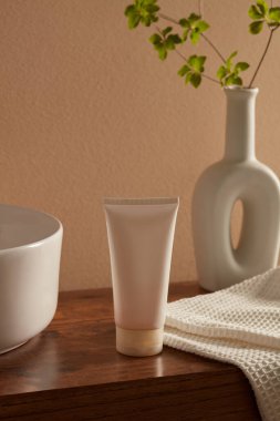Boş beyaz plastik kozmetik tüpü, porselen vazo, yeşil yaprak ve tahta üzerinde pamuk havlu. Krem veya temizleyici için kozmetik maketi. En az konsepte sahip reklam ürünü sahnesi