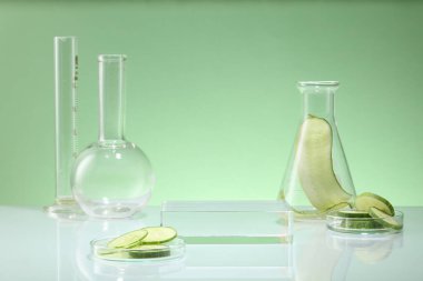 Pastel arka planda salatalık dilimleriyle süslenmiş laboratuvar camları. Salatalık özünün kozmetik ürünlerini sergilemek için boş şeffaf podyum. Laboratuvarda bilimsel araştırma kavramı