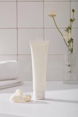 Yüz nemlendirici kremi ya da yüz temizleyicisi, havlu, çiçek vazosu ve beyaz karo arka planda süslenmiş ipek böceği kozaları için etiketsiz bir plastik tüp. Banyo konsepti, günlük rutin içerik