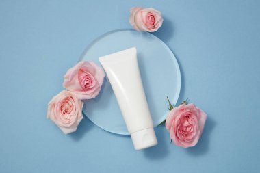 Düz planlı promosyon fotoğrafı, mavi arka planda şeffaf podyum ve güzel güllerle süslenmiş beyaz plastik plastik plastik plastik bir kap. Gül özünün kozmetiği