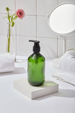 Kare podyumda etiketsiz yeşil bir şişe, banyo içerisindeki beyaz masada pompalanan pompalı gövde bakım ürünü. Beyaz havlular, çiçek tabanı ve beyaz duvar arkaplanındaki ayna..