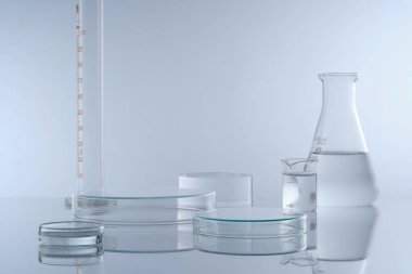 Beyaz zemin üzerinde renksiz sıvı bulunan laboratuvar cam kaplamalı minimal arkaplan. İki petri kabı, kozmetik ürün sunumu için boş platformlar oluşturuyor. Ön görünüm.