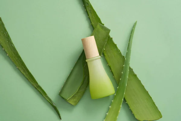 Top view of gradient green bottle with wooden cap and fresh aloe vera leaves decorated on green background. Mockup scene for cosmetics for body and hair care from natural ingredients.