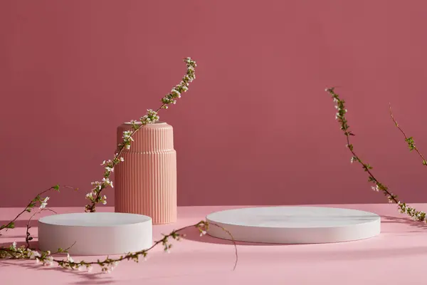 Front view of two white empty podiums and flowers branches on pink background. Pedestal for cosmetic product and packaging mockups display presentation. Minimal concept.
