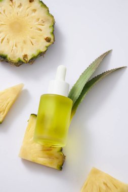 Damla kapaklı sarı sıvı içeren etiketsiz bir cam şişe, taze ananas dilimleri ve beyaz arka planda yapraklar. Serum ürünü için doğal özütten bir model. Üst görünüm.