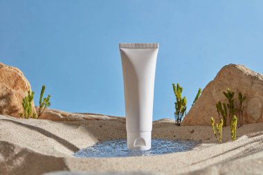 Doğal konsept ile kozmetik ürünleri için model sahnesi. Küçük su birikintisinde etiketsiz beyaz plastik tüp, kum, yeşil ağaçlar ve mavi gökyüzünde kayalar. Ön görünüm, tasarım için alan.