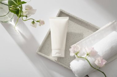 Kare tepside sarılmış havluyla etiketlenmemiş beyaz bir tüp, şeffaf bir vazo. Yüz kremi, temizleyici, vücut losyonu veya şampuan için beyaz kozmetik tüp.