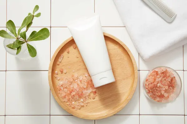 Unlabeled white tube placed on a round wooden dish decorated with a bowl of pink salt, towel and brush. Himalayan pink salt is often used to remove dead skin cells on face or body