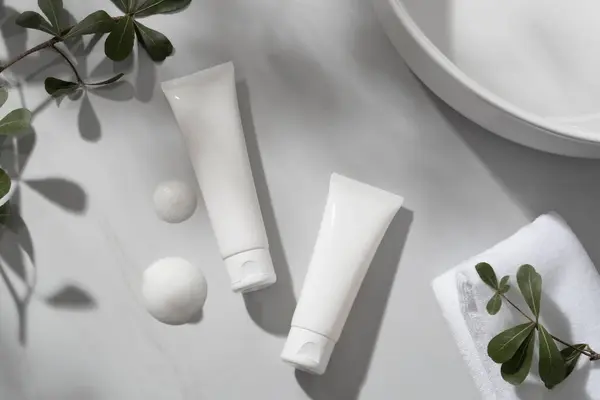 Two empty label tubes decorated with white cleanser foam texture, a towel and some plant branches. Natural soft shadow. Cleansing product advertising.