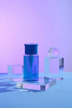 Cam şeffaf dikdörtgen bir podyumun üzerinde duran ve geometrik şekilli nesnelerle sergilenen bir pompa dağıtıcı şişesi. Cilt bakım ürünü modeli