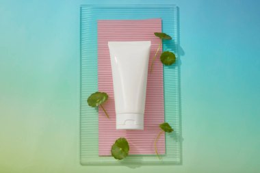 Gradyan mavi arka planda, etiketsiz beyaz bir tüp tırtıklı akrilik çarşaf üzerine konur. Gotu kola (Centella asiatica) çeşitli etkin maddeler içerir