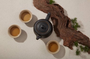 Toprak çaydanlık ve kumlu arka planda sergilenen birkaç çay fincanı içeren düz bir çay seti. Yeşil yapraklarla süslenmiş büyük bir ağaç dalı.