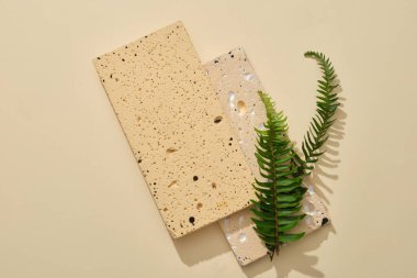 Açık bej arka planda eğreltiotu ile süslenmiş iki taş podyum. Ürün sunumu için podyumda boş alan