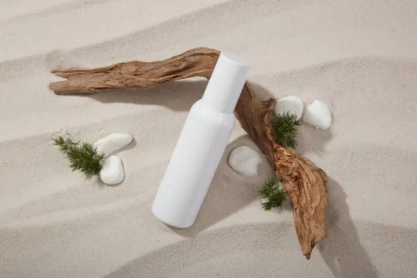 A white bottle leaning on a tree branch decorated with some gravels on the sand. Mockup of beauty cosmetic bottle product with skin care concept