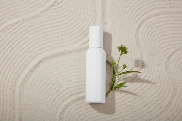 Unbranded cosmetic bottle decorated with a flower branch over a sandy background with waves pattern. Natural organic beauty cosmetics concept