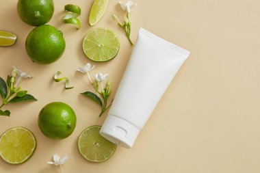 Bej arka planda, bazı Lime (Citrus aurantiifolia) dilimleri, kabukları ve çiçek dalları beyaz bir tüp ile düzenlenir. Boş etiketli bir ürün modeli. Boşluğu sağ tarafa kopyala