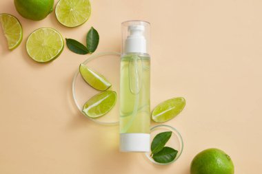 Bej arka planda Lime (Citrus aurantiifolia) dilimleri ve yeşil yapraklarla gösterilen boş etiket pompa şişesi dağıtıcısı. Toner veya serum kozmetik ürün tanıtımı