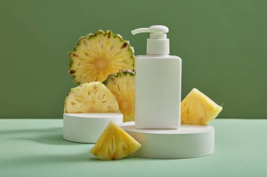 Üzerinde boş etiket şişesi ve yeşil arka planda ananas dilimleri olan beyaz, yuvarlak bir podyum. Ananaslardan elde edilen yatıştırıcı doğal güzellik ürünü kavramı (Ananas comosus)