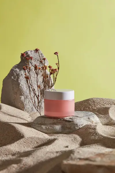 A stone with a pink jar placed on, decorated with flowers and few stones on the sand. Natural skincare beauty product concept