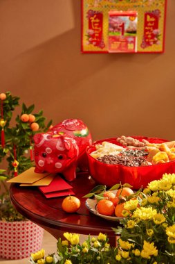 Üzerinde domuz kumbarası, mandalina ve bir kutu kurutulmuş şeker olan bir masa. Sarı çiçek. Takvim kahverengi duvarda asılı..