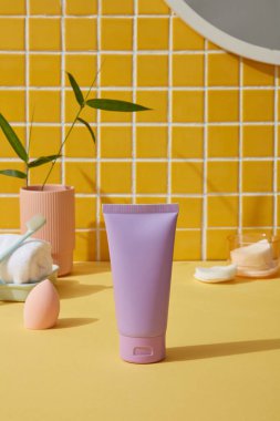 Makyaj süngeri, havlu, pamuk pedi ve sarı banyoda saksı. Ürün reklamında etiket kullanılmayan mor tüp