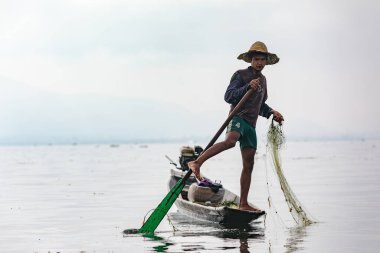 Bir balıkçı Inle Gölü 'nde tek ayağının üzerinde durur ve bir bacağını uzun teknenin dümenine dolayıp ellerini balık yakalamak için serbest bırakır, Myanmar, 12 Nisan 2018