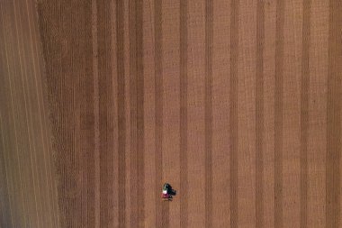 Küçük, yalnız bir traktör. Büyük kahverengi bir tarlada sabanla. Toprak doğrudan yukarıdan görünüyor.