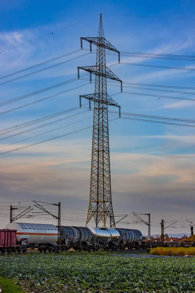 A freight train on a railroad line in front of a high-voltage pylon in a rural area at sunset