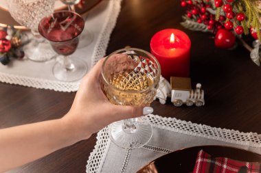 A warm, festive Christmas dinner setting featuring a hand holding a glass of wine. The table is adorned with candles, decorative elements, and lace placemats, creating a cozy and elegant atmosphere. clipart