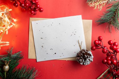 Kırmızı arka planda bir çam kozalağı, kırmızı böğürtlen ve peri ışıklarının yer aldığı, neşeli dekorasyonlarla çevrili boş bir Noel kartı sıcak bir tatil atmosferi yaratıyor..