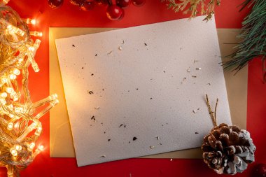 Boş bir Noel kartı, çam kozalağı, köknar dalları ve sıcacık ip ışıkları gibi neşeli elementlerle çevrili kırmızı bir zemin üzerinde duruyor. Sıcak bir tatil atmosferi çağrıştırıyor..