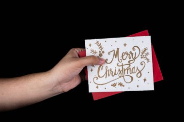 Bir el, Noel ruhunu ve mevsimin sıcaklığını somutlaştıran, siyah arka planda zarif altın harflerle süslenmiş, neşeli bir Noel kartı tutuyor..