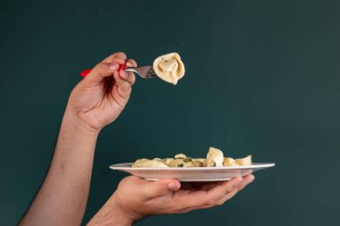 A close-up of a hand holding a fork with tortellini pasta above a plate. The background is green, creating a simple yet elegant culinary scene. clipart