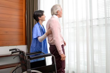 Yaşlı bir adamla ilgilenen Asyalı bir hemşire yaşlılar sağlık merkezinde tekerlekli sandalyeden kalkıyor..