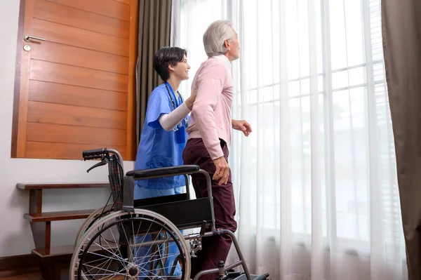 An Asian nurse taking care of an elderly man getting up from wheelchair at senior healthcare center.
