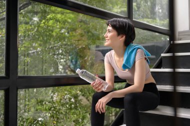 Asyalı genç bir kadın evde egzersiz yaptıktan sonra tatlı su içiyor, sağlıklı yaşam tarzı konsepti.