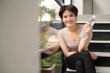 Asyalı genç bir kadın evde egzersiz yaptıktan sonra tatlı su içiyor, sağlıklı yaşam tarzı konsepti.